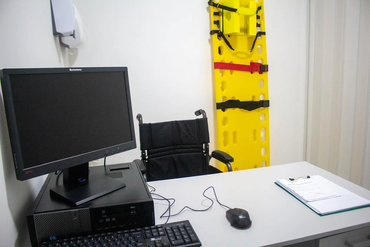Posto de enfermagem com monitor, cadeira de rodas e prancha de imobilização na parede